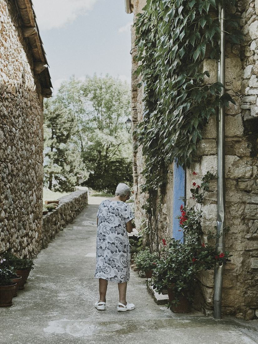 Mujer mayor caminando sola con calma por un callejón de pueblo entre plantas