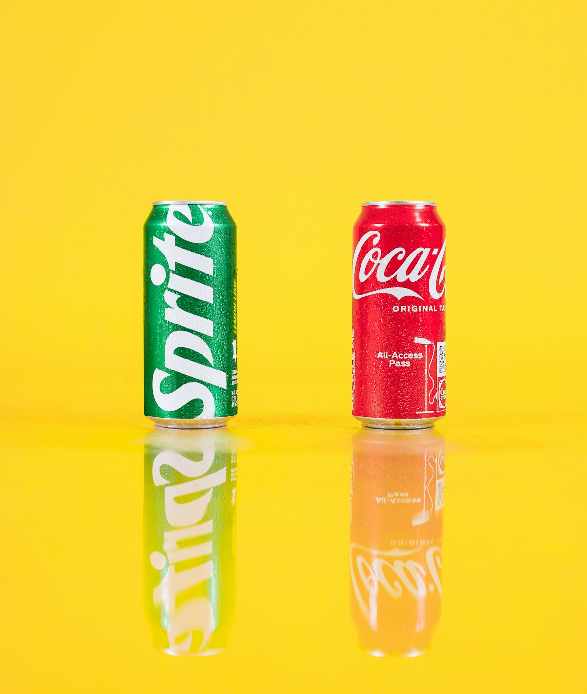 Dos latas de refresco (Sprite y Coca-Cola) sobre un fondo amarillo