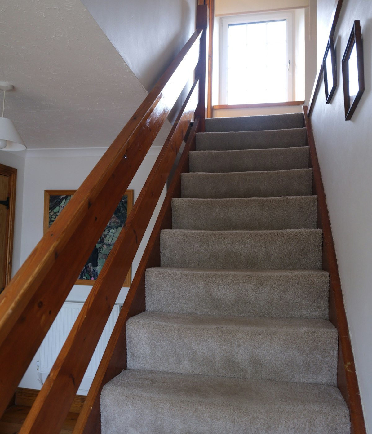Escalera alfombrada de una casa sencilla, con barandal de madera firme e iluminada por luz natural de una ventana