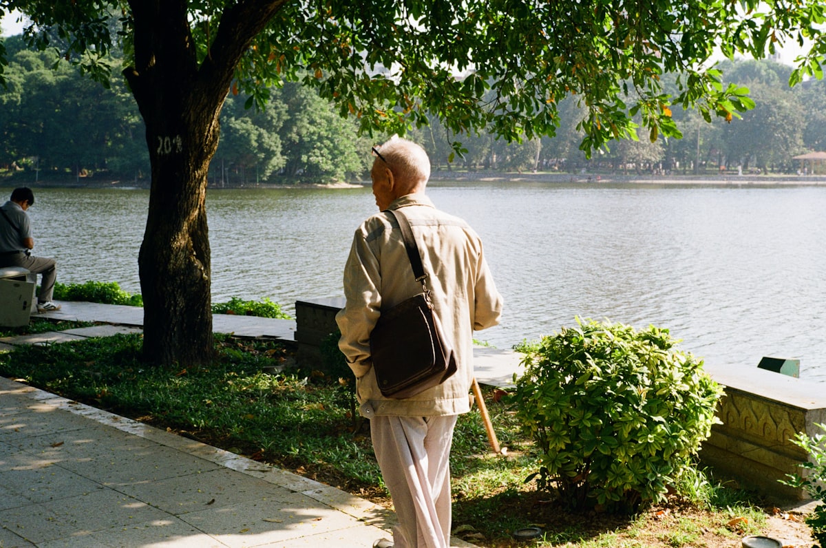 Hombre mayor caminando solo junto a un lago, en un parque arbolado